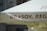 A close-up of a large outdoor umbrella featuring the word 'BRASOV' prominently printed in bold, brown letters on a light-colored fabric. The background consists of a blurred portion of a building and another umbrella underneath.