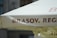 A close-up of a large outdoor umbrella featuring the word 'BRASOV' prominently printed in bold, brown letters on a light-colored fabric. The background consists of a blurred portion of a building and another umbrella underneath.
