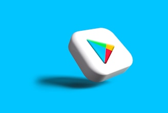 A white 3D cube with rounded edges displaying a colorful triangular logo on its surface, set against a solid bright blue background. The cube casts a subtle shadow, giving a sense of depth.