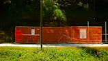 A construction site is enclosed by bright orange mesh fencing, set against a dense, green forest backdrop. The fencing includes several warning signs and notices, while the ground is covered with a gravel path. The sun casts a shadow across part of the scene, creating a vivid contrast between the colors.