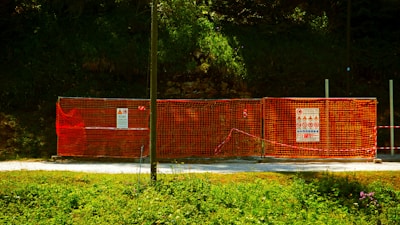 A construction site is enclosed by bright orange mesh fencing, set against a dense, green forest backdrop. The fencing includes several warning signs and notices, while the ground is covered with a gravel path. The sun casts a shadow across part of the scene, creating a vivid contrast between the colors.