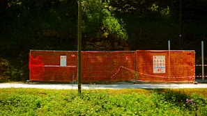 A construction site is enclosed by bright orange mesh fencing, set against a dense, green forest backdrop. The fencing includes several warning signs and notices, while the ground is covered with a gravel path. The sun casts a shadow across part of the scene, creating a vivid contrast between the colors.