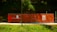 A construction site is enclosed by bright orange mesh fencing, set against a dense, green forest backdrop. The fencing includes several warning signs and notices, while the ground is covered with a gravel path. The sun casts a shadow across part of the scene, creating a vivid contrast between the colors.