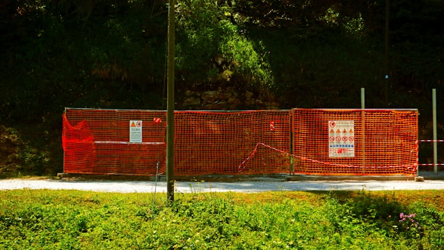 A construction site is enclosed by bright orange mesh fencing, set against a dense, green forest backdrop. The fencing includes several warning signs and notices, while the ground is covered with a gravel path. The sun casts a shadow across part of the scene, creating a vivid contrast between the colors.