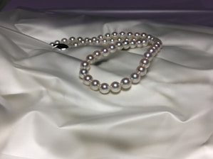 A strand of pearls is elegantly arranged on a smooth, satin-like surface, displaying their luster and reflecting light softly. The pearls are evenly sized and have a classic round shape, enhancing their timeless appeal.