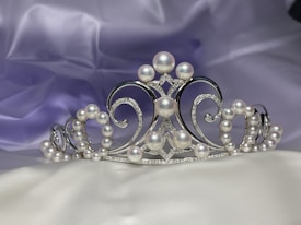 A luxurious silver tiara adorned with pearls and small diamonds rests on a soft, silky fabric backdrop in shades of lavender and white. The tiara features intricate scrollwork with a symmetrical design, combining elegance and regal sophistication.