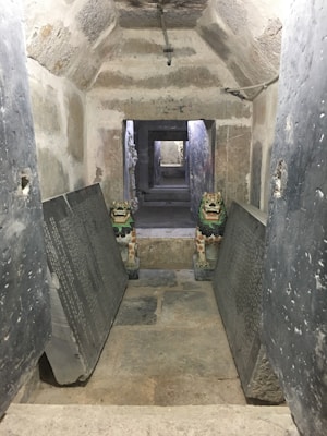 A narrow stone corridor with a series of arched ceilings leads the viewer into deeper passages. Two colorful, ornate statues resembling mythical creatures guard the entrance, placed on either side of the entryway. Large engraved stone tablets are propped against the walls, adding to the historic and ancient ambiance.