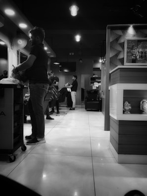 A black and white photo of a modern hair salon with several people engaged in hair cutting and styling activities. The room has contemporary lighting fixtures and decorative elements, including a painting of Buddha and small statues. The salon has a polished tile floor and various salon equipment such as a barber chair and a cart.