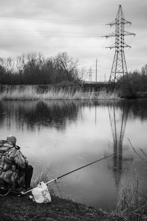 A person wearing a camouflage jacket is fishing by a calm pond. Tall grass lines the edge of the water, and power lines with large metal towers are visible in the background. The sky is overcast, and the scene appears tranquil.