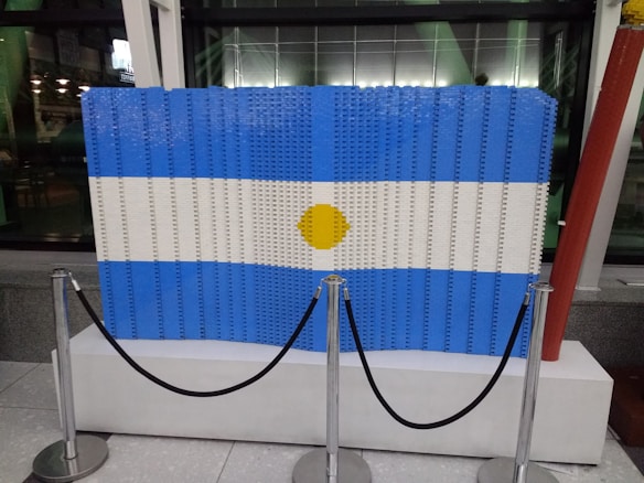 A large rectangular display constructed from blue, white, and yellow LEGO bricks, resembling the flag of Argentina, is showcased on a white platform behind a black velvet rope barrier. The background includes a glass facade and indoor lighting.
