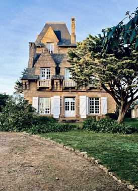 A charming stone house with an ornate roof and chimney, featuring shuttered windows and surrounded by lush greenery and a well-kept lawn.