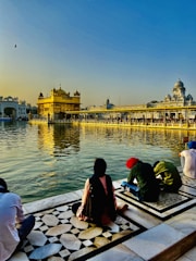 A serene scene by a holy water body reflecting a golden temple in the background, under a clear blue sky with warm sunlight. People are sitting on a patterned marble platform near the water, engaging in prayer or reflection.