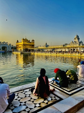 A serene scene by a holy water body reflecting a golden temple in the background, under a clear blue sky with warm sunlight. People are sitting on a patterned marble platform near the water, engaging in prayer or reflection.