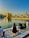 A serene scene by a holy water body reflecting a golden temple in the background, under a clear blue sky with warm sunlight. People are sitting on a patterned marble platform near the water, engaging in prayer or reflection.