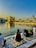 A serene scene by a holy water body reflecting a golden temple in the background, under a clear blue sky with warm sunlight. People are sitting on a patterned marble platform near the water, engaging in prayer or reflection.