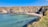 A tranquil cove with turquoise water gently lapping against a small sandy beach. The shoreline is surrounded by steep rocky cliffs with patches of grass and moss. The clear blue sky overhead enhances the peaceful atmosphere of the secluded location.