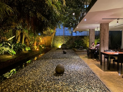 A tranquil interior space featuring a narrow, stone-covered pathway flanked by lush greenery and soft lighting. The setting includes two tables with seated individuals on the right, positioned under a modern ceiling with spotlights. The overall atmosphere suggests a blend of nature with contemporary design elements.