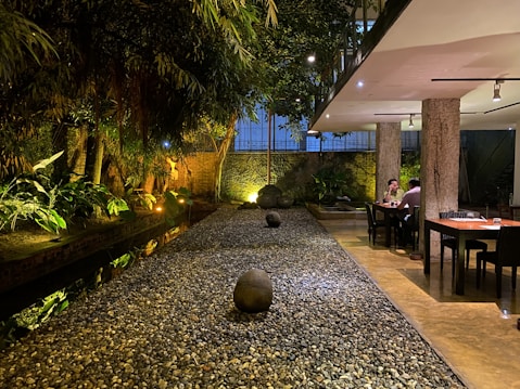 A tranquil interior space featuring a narrow, stone-covered pathway flanked by lush greenery and soft lighting. The setting includes two tables with seated individuals on the right, positioned under a modern ceiling with spotlights. The overall atmosphere suggests a blend of nature with contemporary design elements.