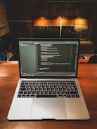 A laptop displaying coding software is placed on a wooden desk in a lecture hall. Behind the laptop, chalkboards can be seen with written equations and notes. The environment suggests an educational or programming context.