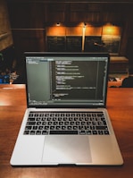 A laptop displaying coding software is placed on a wooden desk in a lecture hall. Behind the laptop, chalkboards can be seen with written equations and notes. The environment suggests an educational or programming context.