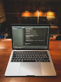 A laptop displaying coding software is placed on a wooden desk in a lecture hall. Behind the laptop, chalkboards can be seen with written equations and notes. The environment suggests an educational or programming context.