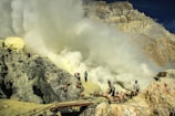Workers are in a rugged, rocky landscape carrying baskets among heavy sulfur smoke near a volcanic site. The scene is industrial and harsh, with people engaged in mining activities.