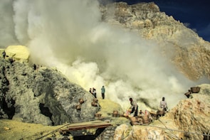 Workers are in a rugged, rocky landscape carrying baskets among heavy sulfur smoke near a volcanic site. The scene is industrial and harsh, with people engaged in mining activities.