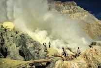 Workers are in a rugged, rocky landscape carrying baskets among heavy sulfur smoke near a volcanic site. The scene is industrial and harsh, with people engaged in mining activities.