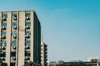 A tall building features numerous air conditioning units mounted on its exterior walls. The building is set against a clear blue sky, with a few other structures visible in the background and some greenery at the bottom.
