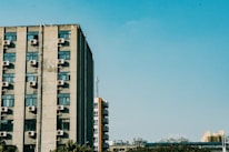A tall building features numerous air conditioning units mounted on its exterior walls. The building is set against a clear blue sky, with a few other structures visible in the background and some greenery at the bottom.