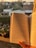 An open book with text in French is held in front of a window, with a sunset illuminating the pages. In the background, there is an outdoor view of residential houses and greenery. A pot of pink flowers sits on a windowsill.