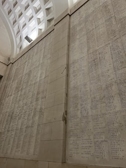 A large stone wall with engraved names and ranks, likely a commemorative or memorial wall. The architecture features an arch with a geometric design on the ceiling, creating a solemn and reverent atmosphere.
