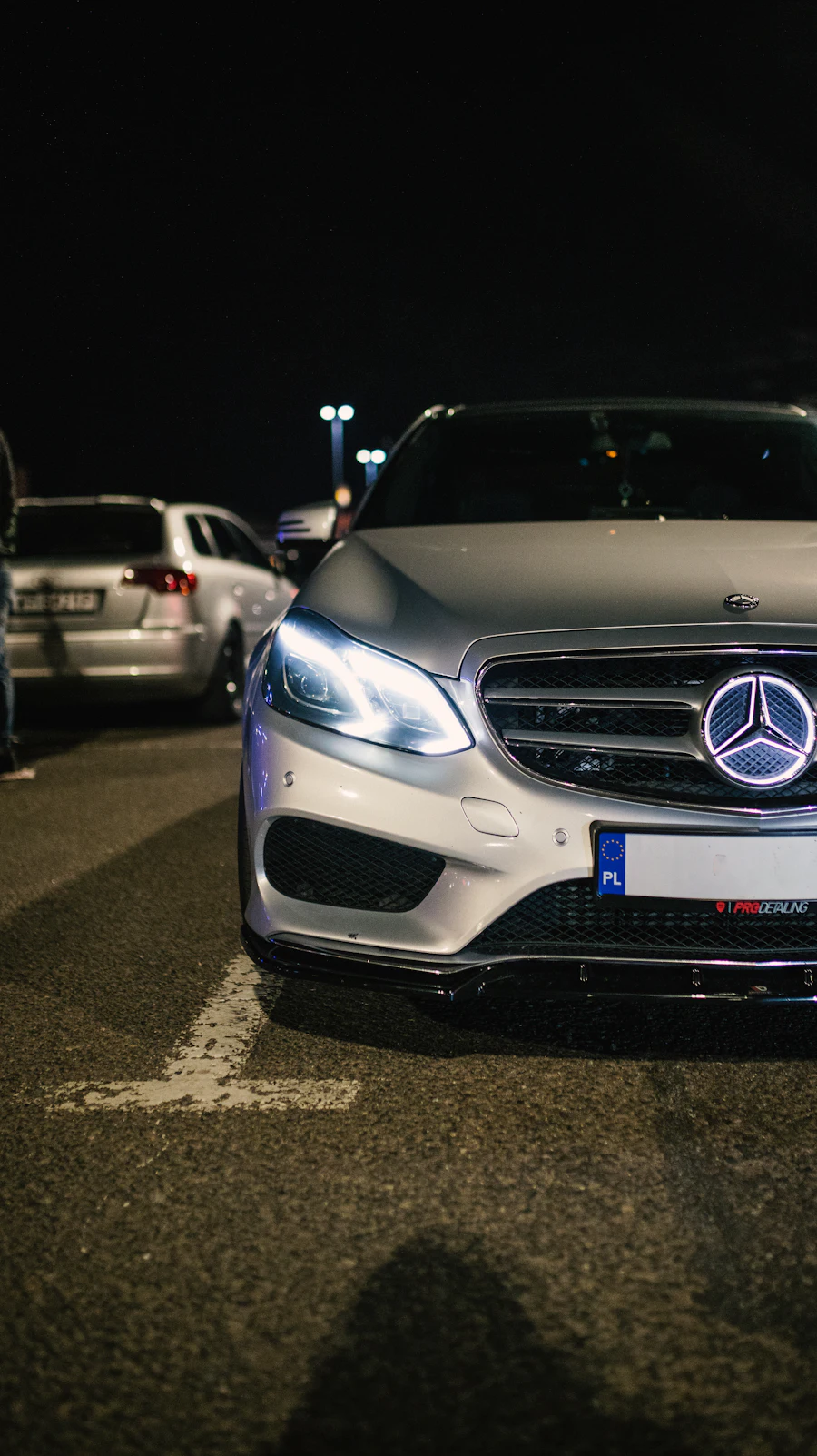 Silver Mercedes E-Class with Polish registration plates, headlights on, parked at night