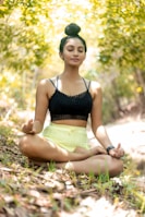 A person is meditating in an outdoor setting, sitting cross-legged on the ground with eyes closed. The surroundings include lush greenery and dappled sunlight creating a peaceful ambiance. The person is wearing a black top and light yellow shorts, with hair tied up in a bun.