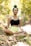 A person is meditating in an outdoor setting, sitting cross-legged on the ground with eyes closed. The surroundings include lush greenery and dappled sunlight creating a peaceful ambiance. The person is wearing a black top and light yellow shorts, with hair tied up in a bun.