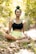 A person is meditating in an outdoor setting, sitting cross-legged on the ground with eyes closed. The surroundings include lush greenery and dappled sunlight creating a peaceful ambiance. The person is wearing a black top and light yellow shorts, with hair tied up in a bun.