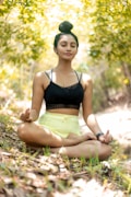 A person is meditating in an outdoor setting, sitting cross-legged on the ground with eyes closed. The surroundings include lush greenery and dappled sunlight creating a peaceful ambiance. The person is wearing a black top and light yellow shorts, with hair tied up in a bun.