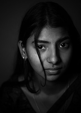 A black and white portrait of a young person looking directly at the camera with a thoughtful expression. The lighting is soft and highlights the person's eyes and facial features. They have long dark hair, with a loose strand falling across the face.