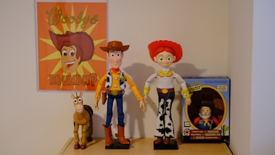 A collection of toys displayed on a shelf, featuring a poster with an illustrated cowboy theme. The central figures include a cowboy doll wearing a brown hat and blue jeans, a doll with red hair and a red hat wearing a rodeo outfit, and a toy horse. To the right is a boxed toy figure that resembles a prospector.