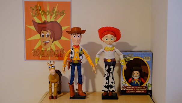 A collection of toys displayed on a shelf, featuring a poster with an illustrated cowboy theme. The central figures include a cowboy doll wearing a brown hat and blue jeans, a doll with red hair and a red hat wearing a rodeo outfit, and a toy horse. To the right is a boxed toy figure that resembles a prospector.