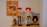 A collection of toys displayed on a shelf, featuring a poster with an illustrated cowboy theme. The central figures include a cowboy doll wearing a brown hat and blue jeans, a doll with red hair and a red hat wearing a rodeo outfit, and a toy horse. To the right is a boxed toy figure that resembles a prospector.