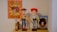 A collection of toys displayed on a shelf, featuring a poster with an illustrated cowboy theme. The central figures include a cowboy doll wearing a brown hat and blue jeans, a doll with red hair and a red hat wearing a rodeo outfit, and a toy horse. To the right is a boxed toy figure that resembles a prospector.