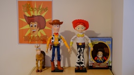 A collection of toys displayed on a shelf, featuring a poster with an illustrated cowboy theme. The central figures include a cowboy doll wearing a brown hat and blue jeans, a doll with red hair and a red hat wearing a rodeo outfit, and a toy horse. To the right is a boxed toy figure that resembles a prospector.