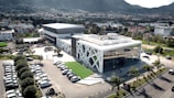 A modern building with distinctive white cross-beam architecture and large glass windows is situated in an urban setting. Surrounding the building is a parking area filled with cars and some landscaped greenery, including palm trees. In the background, a densely populated residential area extends to a mountainous landscape.