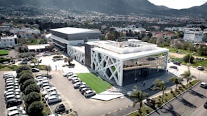 A modern building with distinctive white cross-beam architecture and large glass windows is situated in an urban setting. Surrounding the building is a parking area filled with cars and some landscaped greenery, including palm trees. In the background, a densely populated residential area extends to a mountainous landscape.