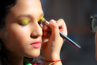 A person is having makeup applied to their face. The makeup artist is using a brush to apply bright green eyeshadow to the person's eyelids. The person has pink lipstick on and a serene expression. The scene is well-lit, highlighting the features of the person's face and the makeup being applied.