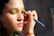 A person is having makeup applied to their face. The makeup artist is using a brush to apply bright green eyeshadow to the person's eyelids. The person has pink lipstick on and a serene expression. The scene is well-lit, highlighting the features of the person's face and the makeup being applied.