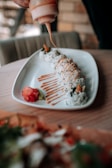 A white rectangular plate holds a row of sushi rolls, drizzled with a creamy orange sauce being applied from a squeeze bottle. Beside the sushi, there is a small amount of pickled ginger and wasabi. The background is blurred, and the setting appears to be a restaurant or dining area.