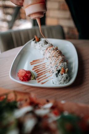 A white rectangular plate holds a row of sushi rolls, drizzled with a creamy orange sauce being applied from a squeeze bottle. Beside the sushi, there is a small amount of pickled ginger and wasabi. The background is blurred, and the setting appears to be a restaurant or dining area.