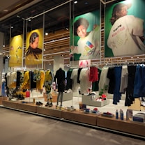 A retail store showcases clothing and sportswear on mannequins and hangers, with large vibrant posters featuring children in sports attire above. The interior displays bright garments and accessories on shelves and racks.
